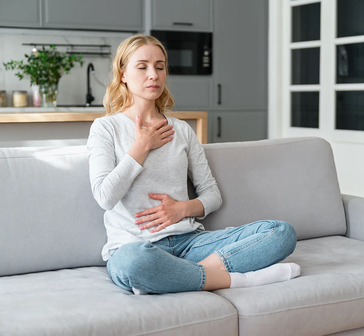 Atemtechnik Zentrale Rolle Eine Frau mit geschlossenen Augen auf der Couch im Schneidersitz sitzend hält sich mit der einen flachen Hand die Brust und mit der anderen den Bauch Bereich. Im Hintergrund ist eine Küche zu sehen