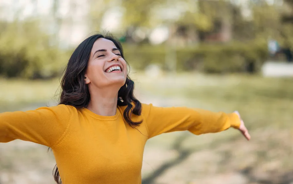 Geringe Nebenwirkungen Eine Frau steht mit ausgebreiteten Armen und geschlossenen Augen lachend auf einer Wiese