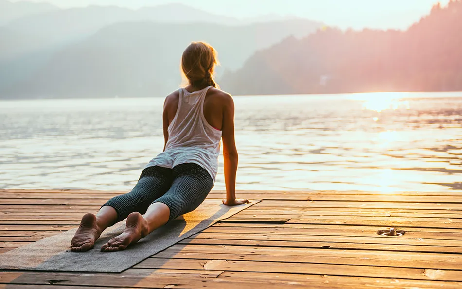 Geringe Pollenbelastung Morgens Abends Eine Frau macht auf einer Matte auf einem Steg vor einem See mit Bergen im Hintergrund Yoga