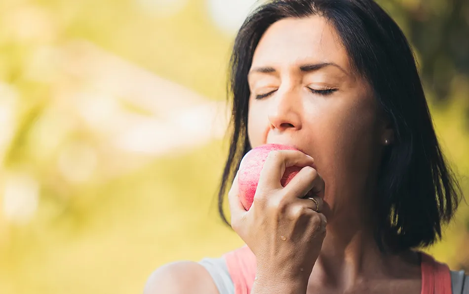 Eine sportlich gekleidete Frau in der Natur beißt mit geschlossenen Augen in einen Apfel