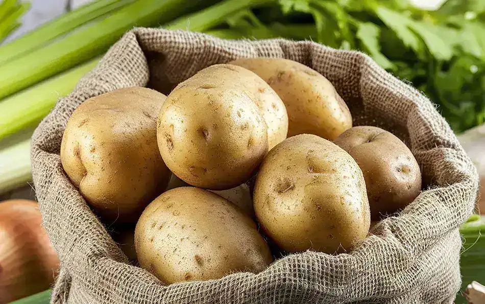 Nahaufnahme von Kartoffeln in einem Sack. Im Hintergrund liegen Staudensellerie und Zwiebeln