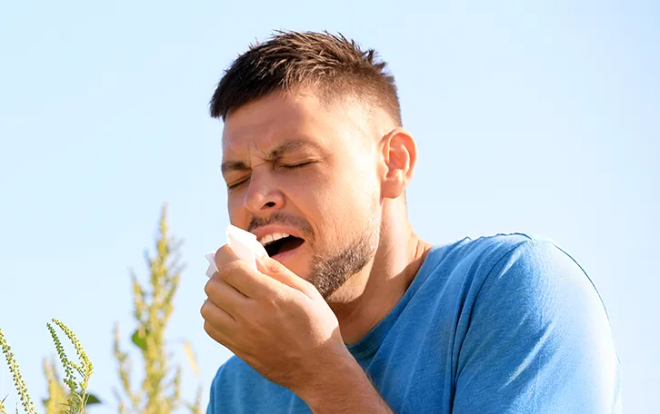 heuschnupfen-symptom-niesen Ein niesender Mann vor blühenden Sträuchern
