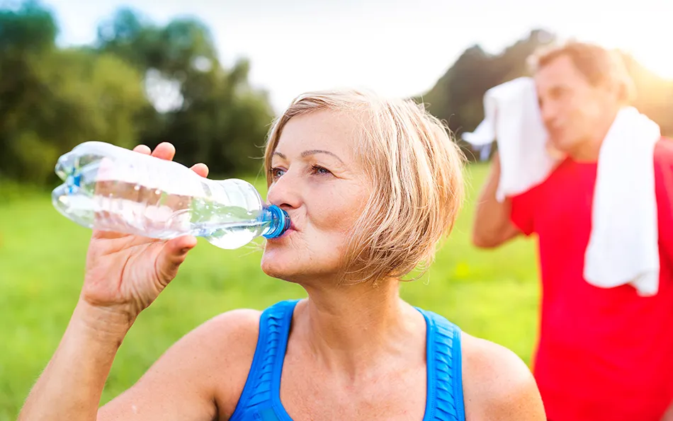 Pausen einplanen Tempo anpassen Eine ältere Frau trinkt aus einer Plastikflasche und ein Mann im Hintergrund trocknet sich mit einem Handtuch den Kopf ab