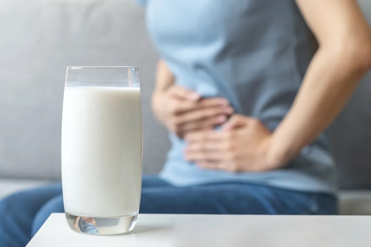 Selbsttest Milchunvertraeglichkeit Im Vordergrund steht ein Glas Milch und im Hintergrund hält sich jemand blau gekeidetes den Bauch-/Darmbereich