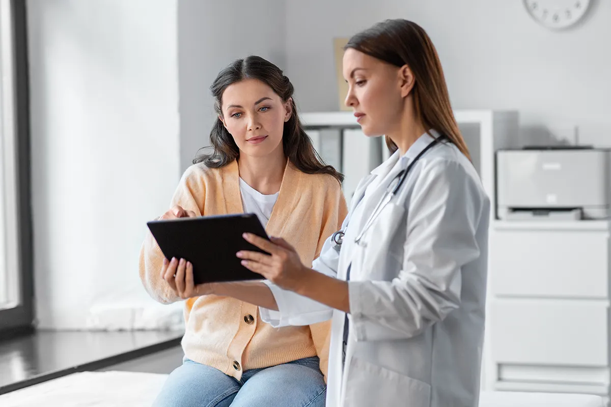 Untersuchung Beratung beim Arzt Eine Frau schaut auf einer Liege sitzend mit einem weiblichen medizinischem Personal mit Stethoskop um den Hals auf ein Pad